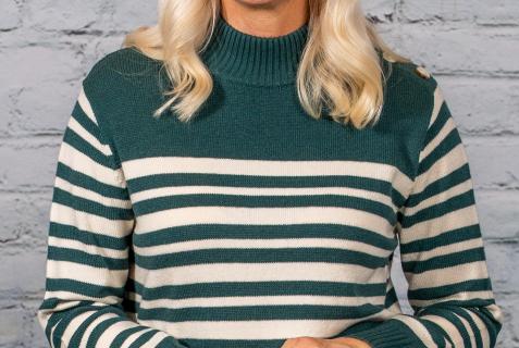 A woman with long, blonde hair smiles while wearing a green and white striped sweater. She stands against a light brick wall, with her hands clasped together in front of her.