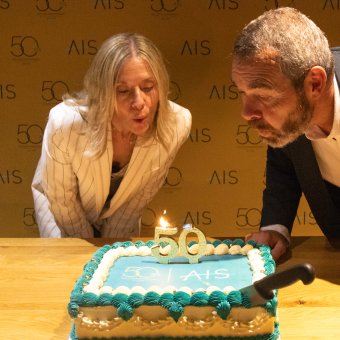 A woman and a man are leaning over a decorated cake with the number "50" on top, preparing to blow out a candle. The background features a banner with the logo "AIS" and the number "50".