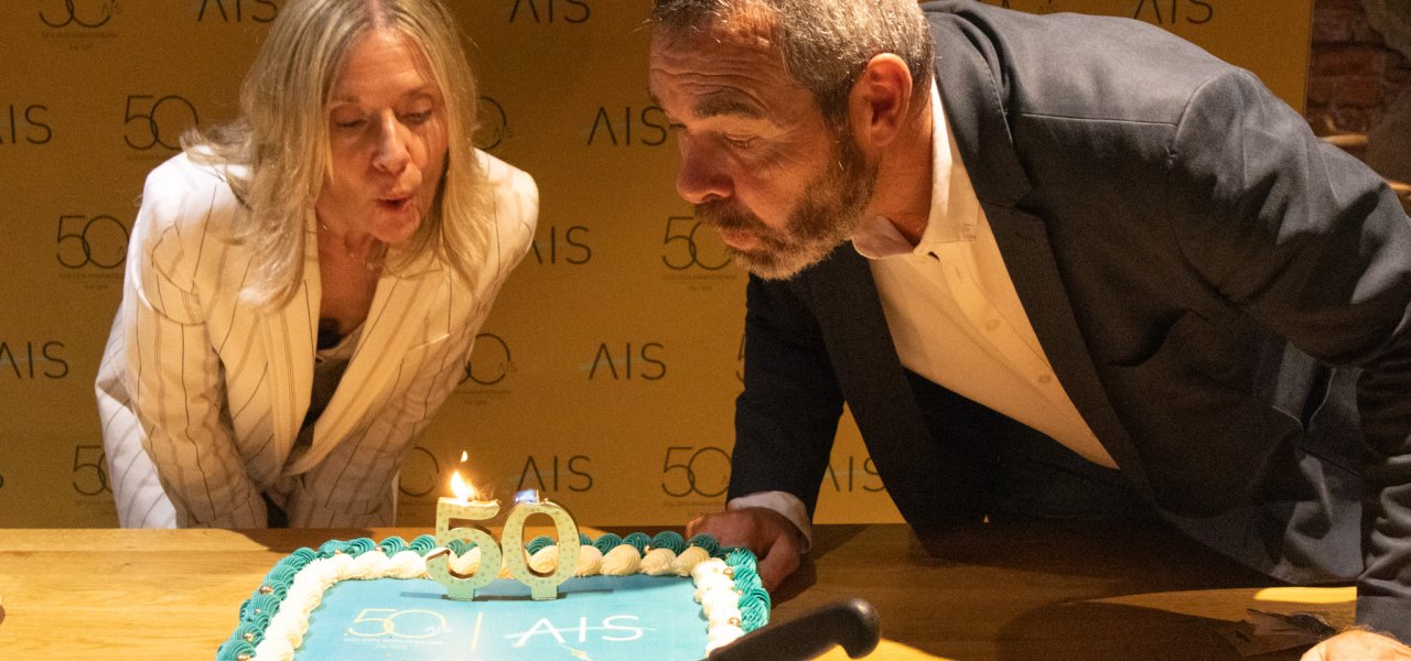 A woman and a man are leaning over a decorated cake with the number "50" on top, preparing to blow out a candle. The background features a banner with the logo "AIS" and the number "50".
