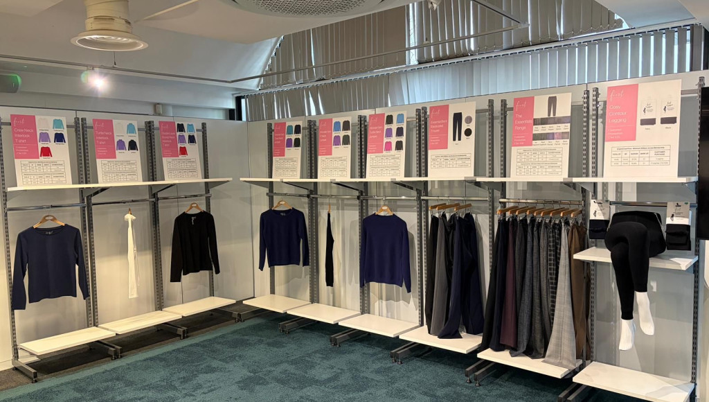 A display featuring clothing samples on racks with various tops and bottoms. Informational boards highlight design details and fabric options. The setting has modern decor with gray carpeting and bright lighting.