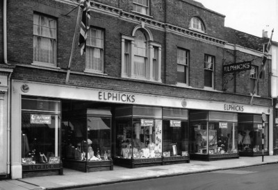 Historic storefront of Elphicks, featuring large display windows showcasing clothing and accessories. An English flag is visible above the entrance, and the building has a traditional architectural style.