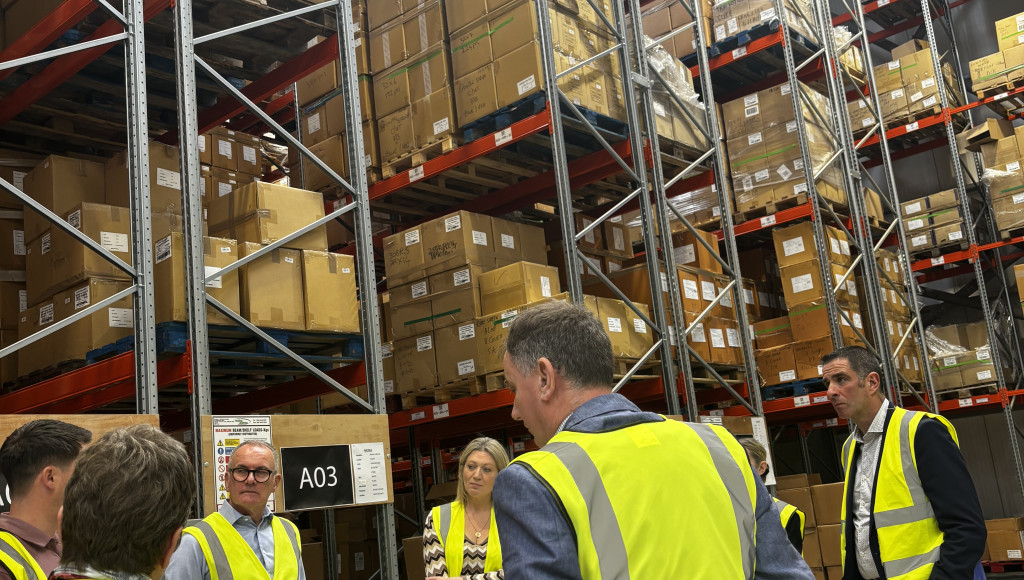 A group of people wearing yellow safety vests stands in a warehouse, discussing while surrounded by tall shelves filled with stacked boxes.