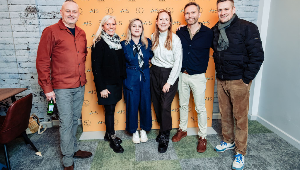 A group of five individuals poses together in front of a bright orange backdrop featuring the text 