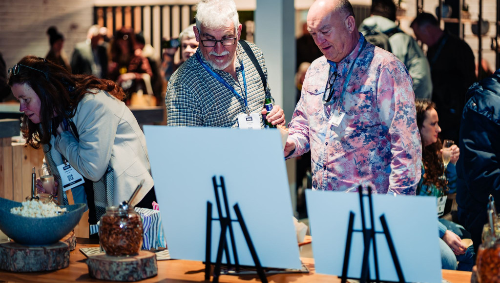 Two men examine blank canvases on easels at an event, surrounded by attendees socializing. A woman looks on in the background, with snacks displayed on a table nearby.