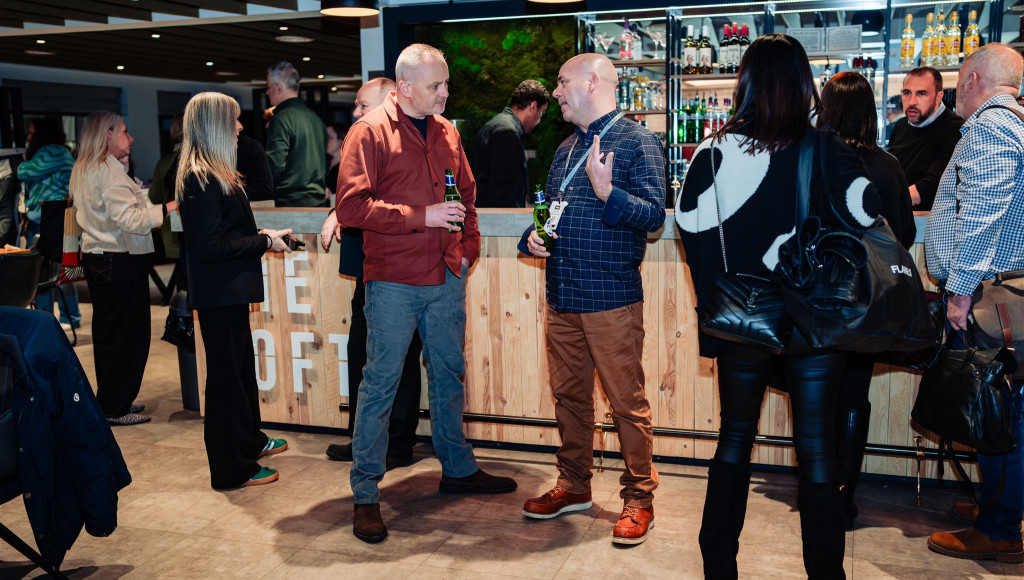 A lively networking event taking place in a modern venue. Two men engage in conversation near a bar, while other attendees socialize in the background. Stylish decor and ambient lighting enhance the atmosphere.