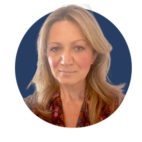 A person with long, wavy blonde hair, wearing a dark floral blouse, is smiling softly against a solid blue background.