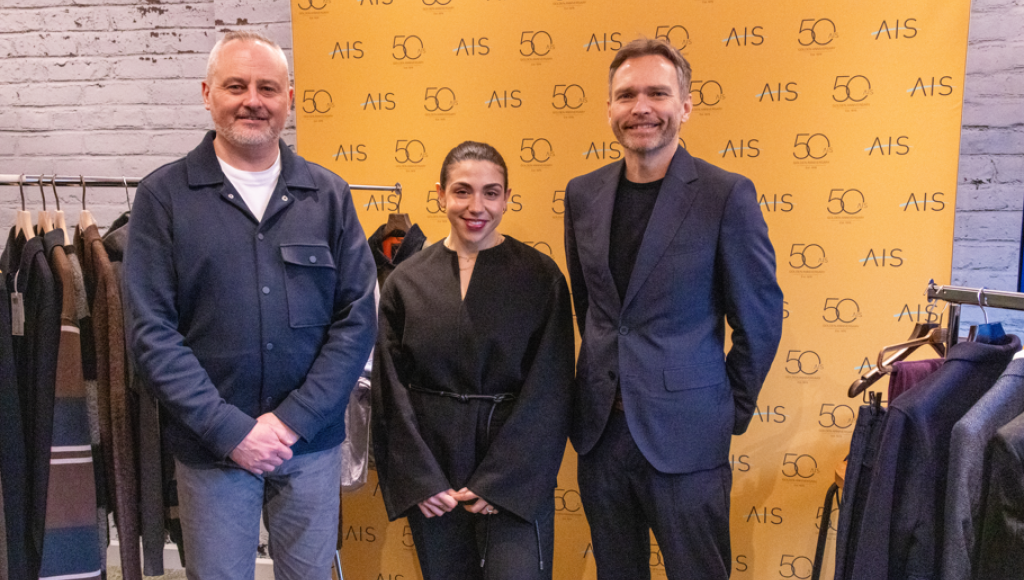 Three individuals pose together in front of a yellow backdrop featuring 