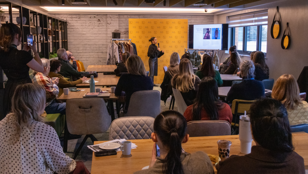 A speaker presents in front of an audience during a seminar, with a screen displaying a slideshow. Attendees sit at tables, engaged and taking notes, while some use their phones to capture the moment. The room features a modern design with bright accents and clothing racks in the background.