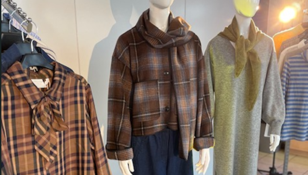 Three mannequins display stylish outfits. The left mannequin features a brown plaid shirt with a bow tie, while the central mannequin showcases a brown plaid cropped jacket with a scarf. The right mannequin wears a gray dress with a tied neck detail.