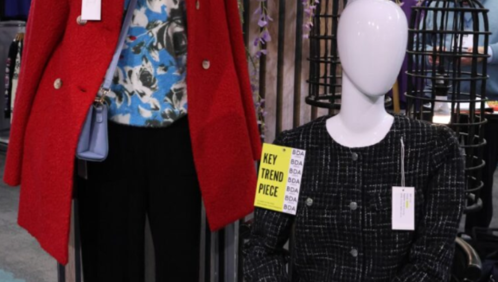 Two mannequins display fashionable outfits. One is wearing a vibrant red coat over a blue floral top, while the other is dressed in a black and white tweed ensemble with large buttons. A sign labeled 