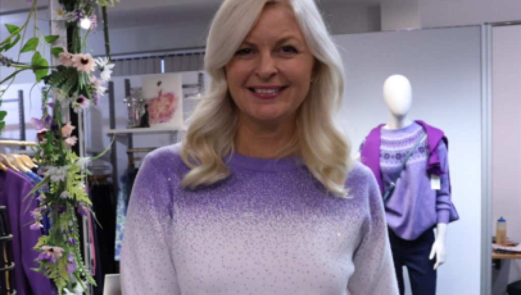 A woman with long, wavy blonde hair smiles, wearing a light purple sweater with a gradient design. She stands in a stylish retail environment with clothing display racks and a mannequin in the background, featuring more purple attire.