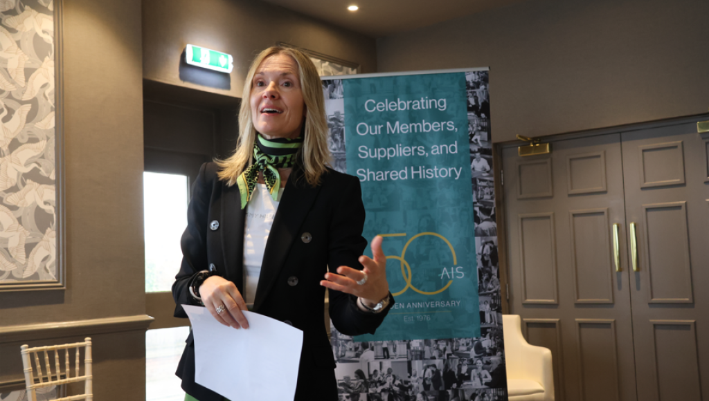 A woman with long blonde hair speaks animatedly while holding a piece of paper. She is wearing a black blazer and a green scarf. Behind her is a banner celebrating an organization's 50th anniversary, featuring text about members, suppliers, and shared history, along with a collage of images.