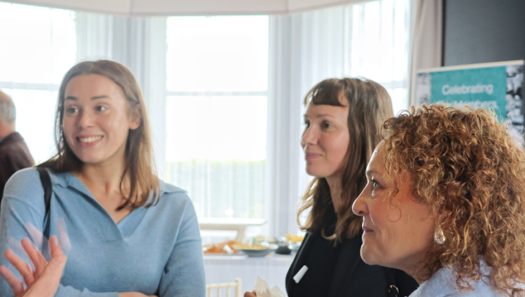 Three women engaged in conversation at a social event. They are smiling and appear to be enjoying the interaction. In the background, there are tables with refreshments.