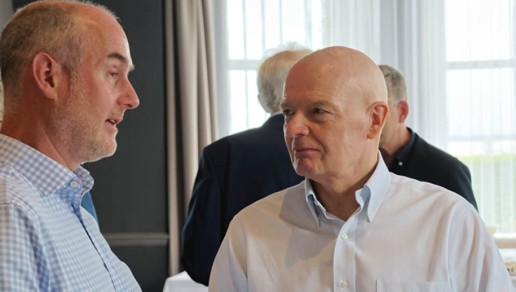 Two men are engaged in conversation indoors, with soft lighting and blurred people in the background. One man is wearing a light blue collared shirt, while the other is dressed in a checkered shirt. Both appear to be listening intently.