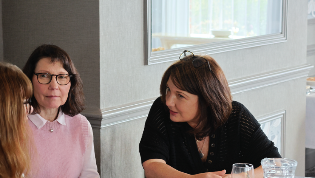 Two women sit at a table engaged in conversation, with a third woman partially visible. They are in a well-lit dining area, featuring elegant decor. One woman wears glasses and a pink sweater, while the other has dark hair and a black top. A glass of water and a pitcher are on the table.