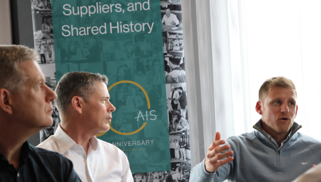 A group of three men engaged in conversation at a gathering, with one man animatedly speaking and gesturing. In the background, a banner highlights the themes of membership, suppliers, and shared history, celebrating an anniversary. Natural light filters through a window.