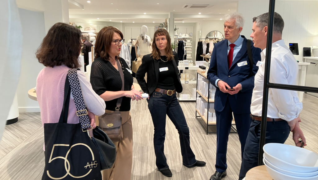 A group of five people engaged in discussion inside a modern retail space, showcasing a variety of clothing and home goods. One woman stands in the foreground speaking, while the others listen attentively. The atmosphere appears collaborative and professional.