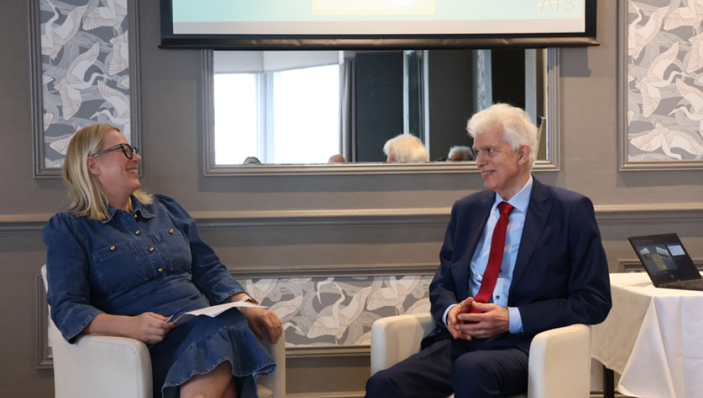 Two people seated in a conference setting. One is a woman with glasses wearing a denim dress, holding a notepad, and the other is a man in a suit and red tie. They appear to be engaged in a conversation, smiling, with a screen displaying the text 