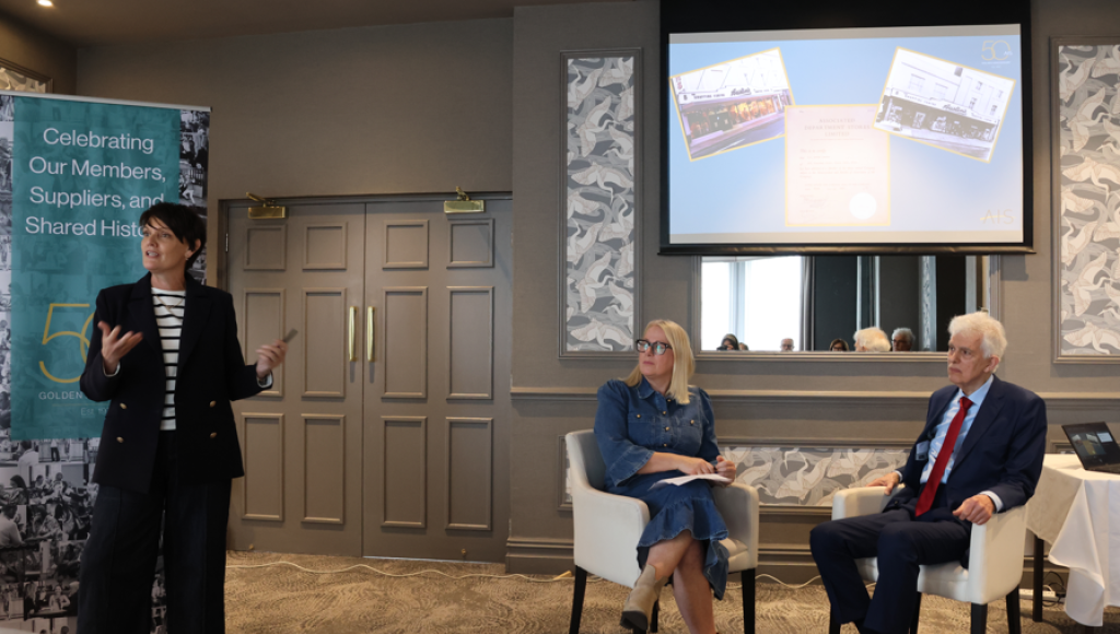 A woman in a black blazer and striped shirt speaks while gesturing. Two seated attendees listen, one wearing glasses and a blue dress, the other in a red tie and suit. A screen displays presentation slides in the background. A banner promotes celebrating members and shared history.