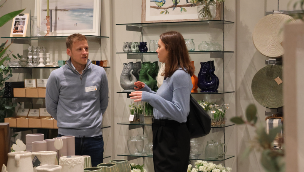 A man and a woman are engaged in conversation in a store filled with decorative items. The shelves display various vases, ceramics, and decorative objects. The woman is wearing a light blue sweater and black pants, while the man is dressed in a light blue pullover.