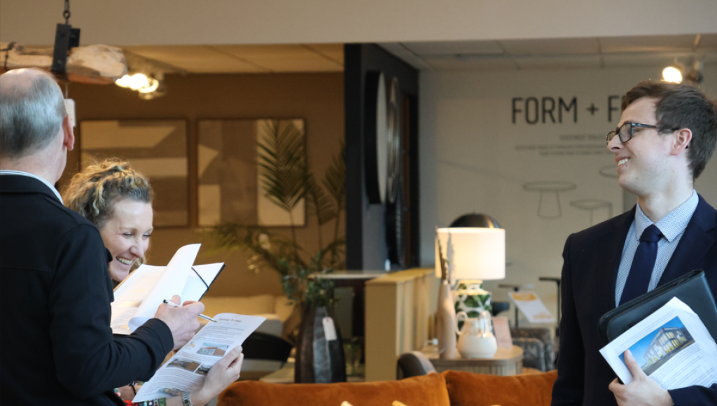 A group of four people engages in conversation in a stylish furniture showroom. One person holds a clipboard while another laughs. A man in a suit looks on, smiling. The background features modern decor and a cozy seating area.
