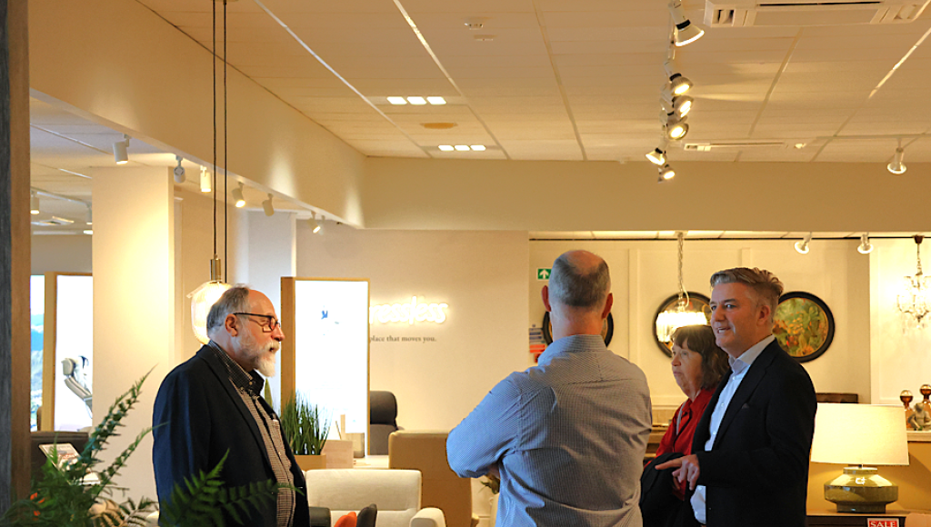 A group of four people engaged in conversation in a well-lit home furnishings store. Two men are facing each other, while a woman and another man stand nearby, observing. Modern furniture and decor can be seen in the background.