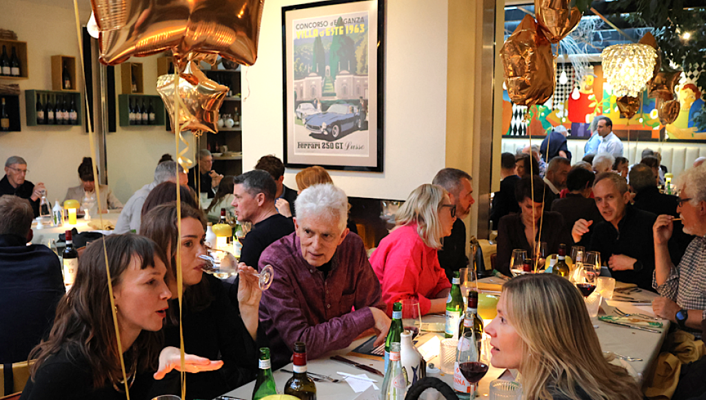 A lively restaurant scene with diners engaged in conversation, some holding drinks. Balloons with a star shape are seen in the foreground, and colorful artwork decorates the walls. The ambiance is warm and celebratory, with several tables occupied by guests.