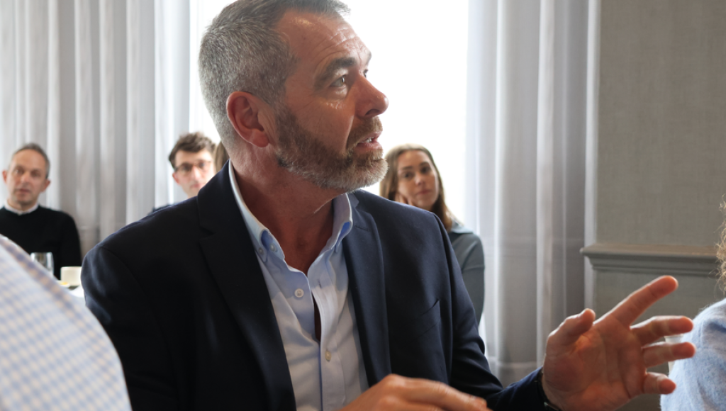 A man with a beard in a blazer gestures while speaking during a discussion at a table. People are seated in the background, listening attentively. Natural light filters through large windows.