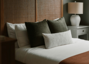 A neatly made bed with a wooden headboard features three decorative pillows in various shades of green and white, alongside a soft, textured throw pillow. A bedside lamp and a nightstand are visible in the background.