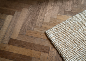 A close-up of a wooden herringbone patterned floor featuring varying shades of brown, complemented by a textured light-colored woven rug at the edge.