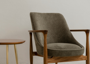 A modern, olive-colored armchair with wooden arms sits next to a small round side table with slender golden legs against a light-colored wall.
