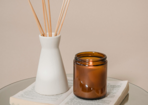 A minimalist arrangement features a white ceramic vase with bamboo reed sticks and an amber glass candle jar resting on an open book, set against a soft beige background.