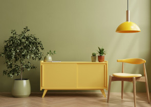 A cozy interior scene featuring a bright yellow cabinet against a green wall, complemented by a yellow pendant light. A potted plant sits beside the cabinet, while another plant is on top. A wooden chair with a minimalist design completes the warm, inviting atmosphere.