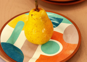 A vibrant yellow pear sits on a colorful, patterned plate with abstract shapes. Two additional patterned plates are visible in the background, resting on a wooden surface.