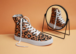 High-top sneaker with a leopard print design and white laces, placed next to a circular mirror reflecting the shoe, against a peach-colored background.