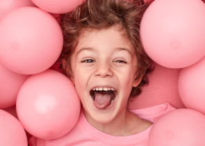 A joyful child with curly hair lying among pink balloons, smiling widely and showing excitement.