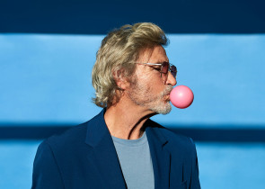A man with gray hair and sunglasses blows a pink bubblegum bubble while wearing a blue blazer over a gray shirt. He stands against a blue background with shadows, showcasing a casual yet stylish demeanor.