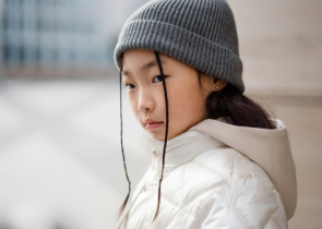 A young girl with long, dark hair wearing a gray beanie and a white puffer jacket looks thoughtfully to the side. Her expression is calm and serious against a blurred urban background.