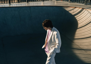 A person wearing a white suit walks alone in a skatepark bowl, with soft shadows creating lines on the surface. Their head is slightly down, and they're positioned toward the edge of the bowl.