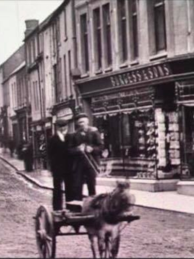 Two men stand beside a donkey cart on a cobblestone street. Behind them, a shopfront displays the name 